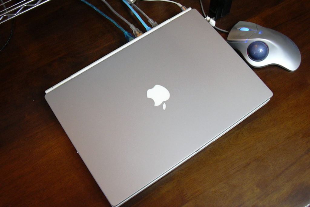 Apple ComputerのノートブックPCであるPowerBook G4 Titaniumを撮影した写真。 木製の机の上に蓋が閉じられた状態で置かれている。 右横にはトラック ボール マウスがある。