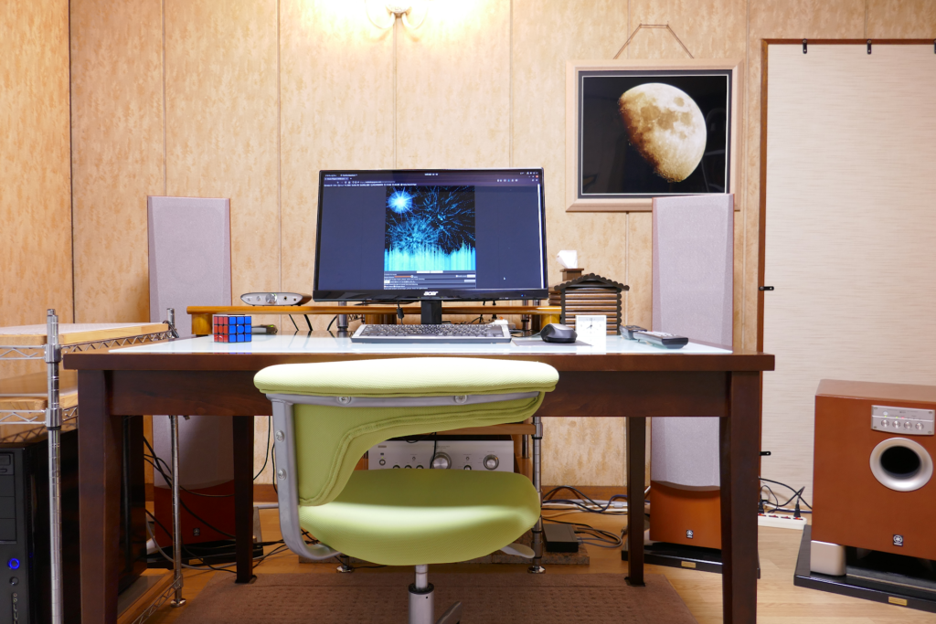 部屋の写真。 中央にはPCディスプレイとキーボードとマウスとオーディオ装置用リモコンが置かれたテーブルと黄緑色の回転椅子がある。 テーブルの左にミドル タワー型の黒いPCがある。 奥にはフロント チャネル用の大きなトールボーイ型スピーカー システムが2本あり、右端には大きなサブウーファーが置かれている。 PCディスプレイには自作の音楽再生プログラムの画面が表示されている。