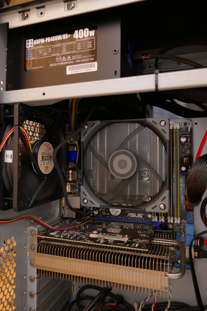 自作PCの内部の写真。 左方にケース ファンがある。 下方にファンレス ヴィデオ カードがある。 中央にファンが回転しているCPUクーラーがある。 クーラーの右横に黄金色のヒート スプレッダーが付いたメイン メモリーが2枚挿入されている。 奥にマザーボードがある。 左上に電源装置の筐体がある。