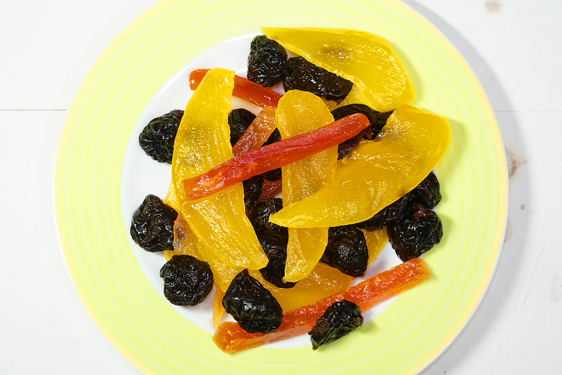 黄緑色の縁取りの丸い皿の上に盛られた水に濡れたドライ フルーツを撮影した写真。 黒色で皺だらけのプルーンと黄色の細長く薄切りにされたマンゴーと赤色の千切りのパパイアが乱雑に盛られている。
