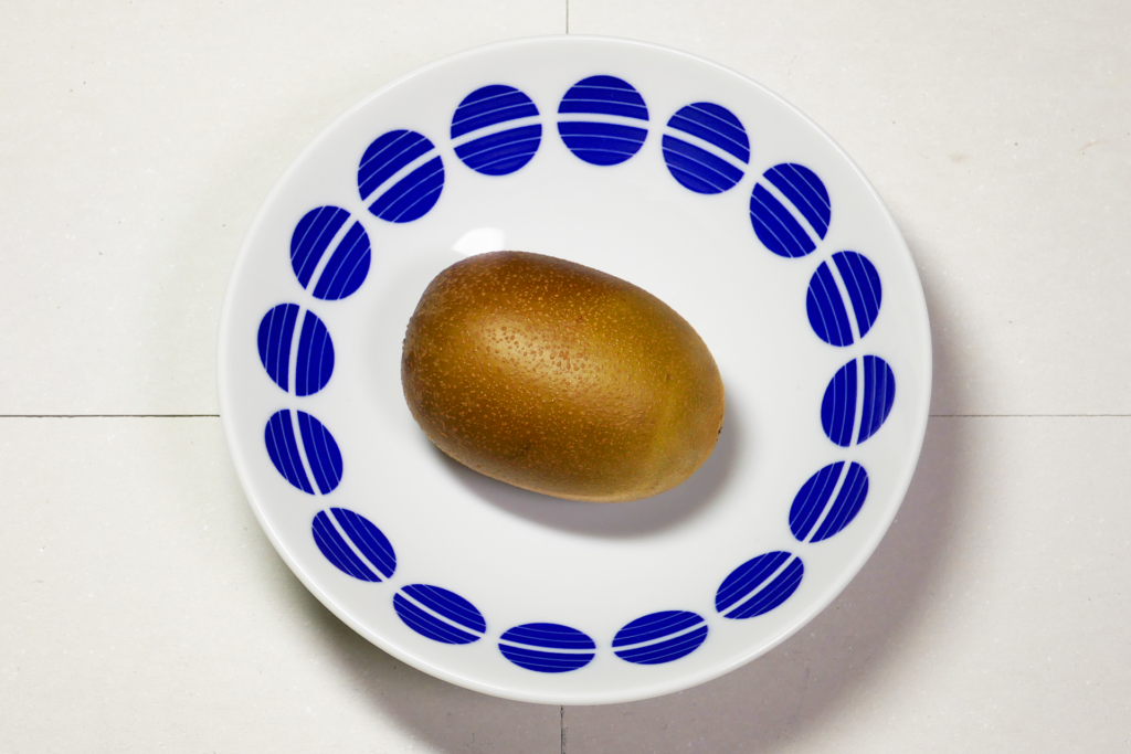 青色の模様が描かれた白色の丸い皿に乗った1個のキウィフルーツを斜め上から撮影した写真。