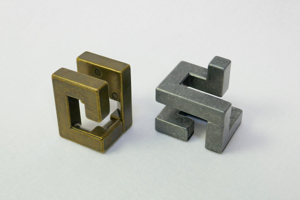 非鏡面の金属製の立体パズルであるHuzzleを撮影した写真。 2つのピースが分離されている。 白色のフェルトの上に置かれている。