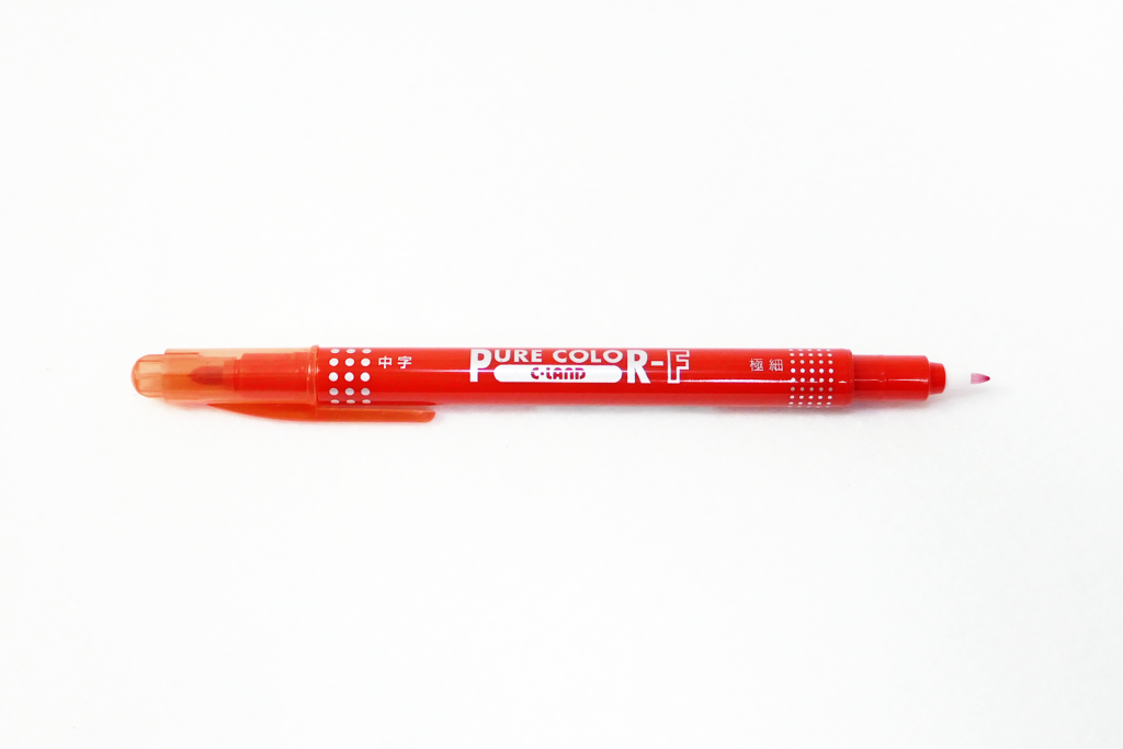 赤色インクのペンを撮影した写真。 ペンのデザインは鮮やかな赤色である。 横向きに置かれている。 向かって左側が中字用で半透明のキャップが付いており、右側が細字用でキャップが外されている。