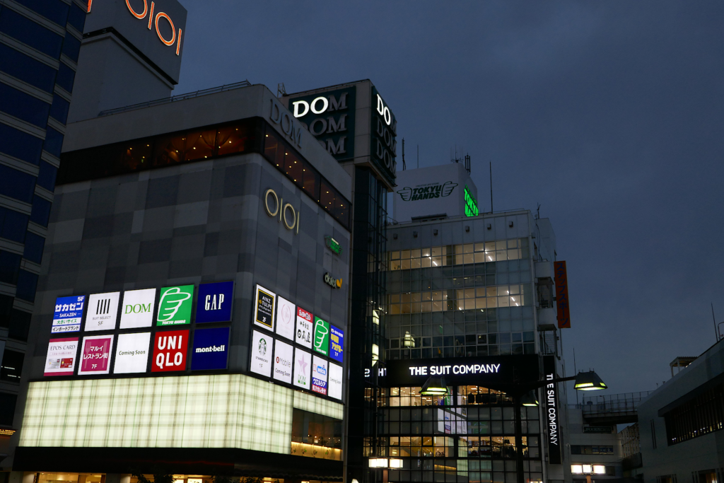 埼玉県さいたま市大宮駅近辺の都市の風景写真。 薄暗い日没時の高層ビルディングが写っている。 商業ビルディングの壁面には入居店舗の光り輝く看板が多数並んでいる。