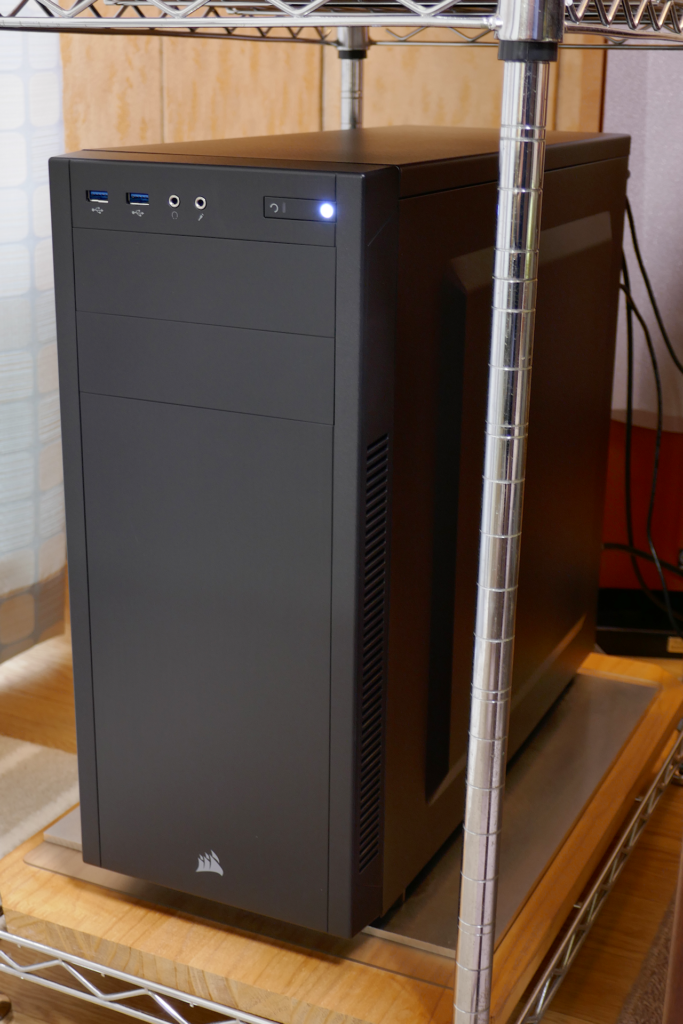 自作PCであるCORSAIR Carbide Series 100R Silentの写真。 テーブルの左横の棚の最下段に設置されている。 ケース全体が黒色で質素なデザインである。 電源ボタンの部分が白色の光を放っている。