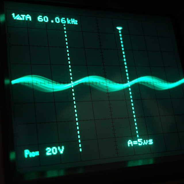オシロスコープの画面の写真。 正弦波のノイズ波形が映し出されている。 周波数は凡そ60kHzとなっている。