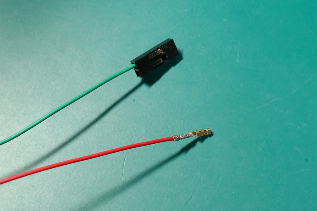 QIコネクターのコンタクト ピンにハンダ付けしてある赤色の被覆線と、QIコネクターのハウジングに差し込まれたコンタクト ピンの付いた緑色の被覆線の写真。